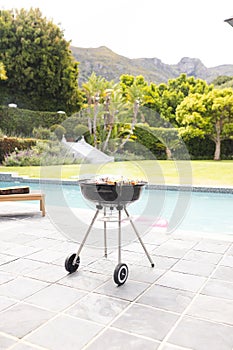 A charcoal grill is ready with food on it, beside a pool in a lush garden with copy space