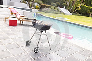 A charcoal grill is smoking beside a pool with a lounge chair and pink float