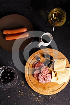 Charcuterie, grilled sausages, cheese, salami, plum olives and a glass of wine on a dark table