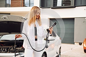 With charging cable in hands. Young woman in white clothes is with her electric car at daytime