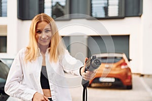 With charging cable in hands. Young woman in white clothes is with her electric car at daytime