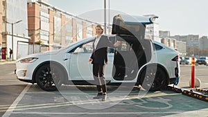 Charging electric car. Charming young woman in business suit dancing joyfully outdoors while her luxury electro car