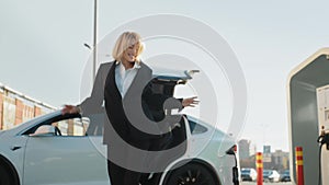 Charging electric car. Charming young woman in business suit dancing joyfully outdoors while her luxury electro car