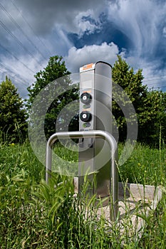 Charging Station For Electric Cars In Green Meadow In Front Of Trees And Electricity Cables
