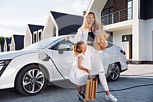 Charging the vehicle together. Young woman with her little daughter is with their electric car outdoors