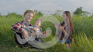 Charismatic child boy is learning to play guitar while mom and dad laugh and admire while relaxing on family picnic in