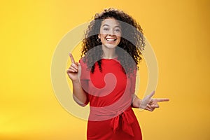 Charismatic cute and happy young curly-haired european female in red dress dancing and pointing up and right, showing