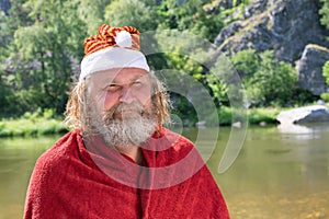Charismatic elderly man with a beard in Santa Claus hat and red coat smiling. Against background of a summer landscape