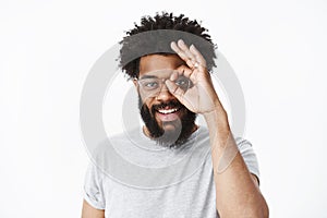 Charismatic friendly african american man with beard, afro hairstyle and pierced nose showing okay gesture over eye and