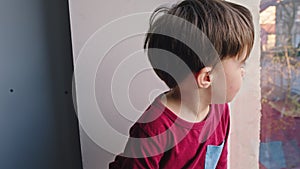 Charismatic small cute boy looking sad through the window he stay at home in quarantine and waiting to go outside to