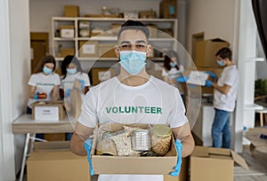 Charity center. Arab activist male volunteer holding food donation box and looking at camera, wearing medical mask