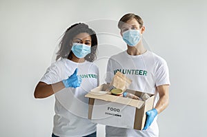 Charity center. Millennial diverse volunteers in face masks holding food donations box and lady showing thumb up