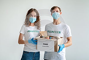 Charity center. Millennial volunteers holding food donations box with products and canned food, wearing medical masks