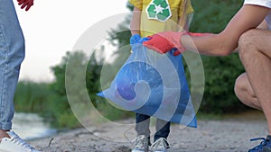 Charity cleaning, young family with little kid in rubber gloves collects refuse in garbage bag on dirty beach from