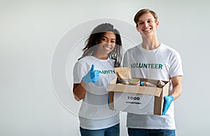 Charity organization. Mixed race volunteers holding food donations box, woman showing thumb up over light wall