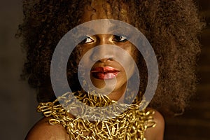 Charming african girl, with black curly hair and gold makeup, accessories,looking away on the dark background, in studio, close up