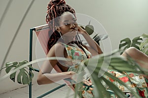 Charming African woman, wearing a multicolor ornament dress, sitting in a hammock chair surrounded by tropical plants. Resting in