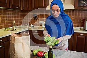 Charming Arab Muslim woman in hijab, holding leaves of fresh salad while unpacking grocery shopping bag in the kitchen