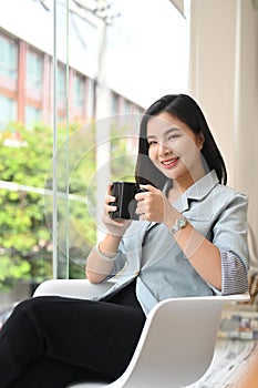 Charming asian businesswoman sitting on armchair, enjoying her coffee break at personal office