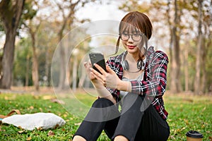 Charming Asian female college student uses her phone and relaxes while sitting on grass in a park