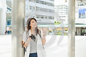 Charming Asian woman in gray shirt and open shirt carrying a backpack and holding camera, make a finger point on the air  from