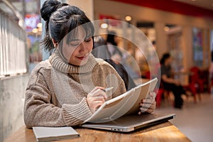 A charming Asian woman is listing her ideas in a book, working remotely at a coffee shop