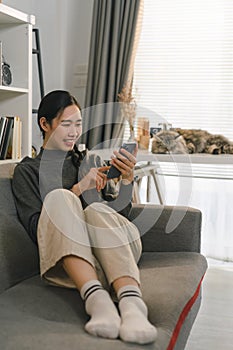 Charming Asian woman is using her smartphone while relaxing on the sofa at home.