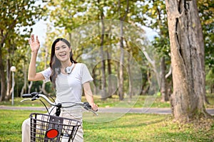 Charming Asian woman waving her hand, greeting her friends while she is on her bike in the park