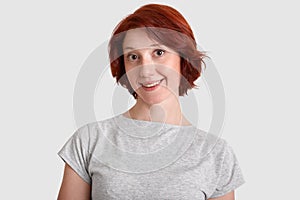 Charming attactive woman with short hairstyle, has broad smile, dressed in casual grey t shirt, isolated over white background.