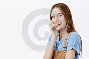 Charming authentic and enthusiastic charismatic young woman in t-shirt and overalls standing half-turned with flirty