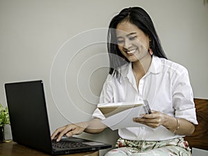 Charming beautiful freelancer in coffee shop holding a book in hand, typing on laptop keyboard with smiling face