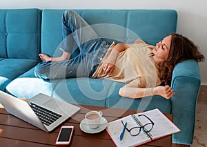 Charming beautiful young girl in glasses with curly hair fell asleep on a blue sofa at home in front of a laptop, notepad and a