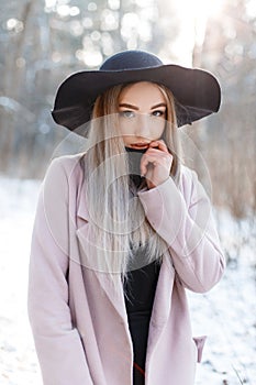 Charming beautiful young woman with blond hair in a knitted dress in a chic black hat in a pink elegant coat