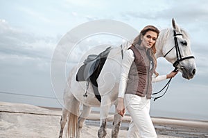 Charming and beautiful young woman wearing stylish jockey outfit is holding the reins and posing with the white horse