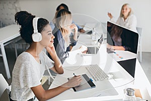 Charming black web-developer listening music in big headphones and using tablet. Indoor portrait of programmers working