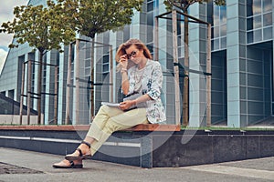 Charming blonde female in modern clothes, studying with a book, sitting on a bench in the park against a skyscraper.