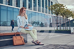 Charming blonde female in modern clothes, studying with a book, sitting on a bench in the park against a skyscraper.