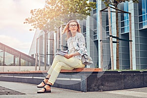 Charming blonde female in modern clothes, studying with a book, sitting on a bench in the park against a skyscraper.