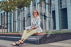 Charming blonde female in modern clothes, studying with a book, sitting on a bench in the park against a skyscraper.