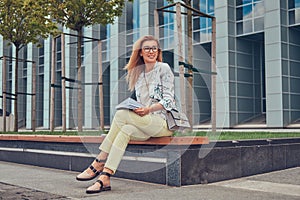 Charming blonde female in modern clothes, studying with a book, sitting on a bench in the park against a skyscraper.