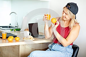 Charming blonde is having breakfast in her kitchen, comparing color of freshly squeezed orange juice and orange itself.