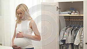 A charming blonde is standing to the left of the opened wardrobe with babyâ€™s clothes. She holds her hands on the belly,