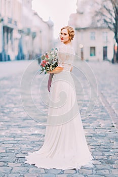 Charming bride in long lace dress holding vintage bouquet looking over shoulder into the camera with old city