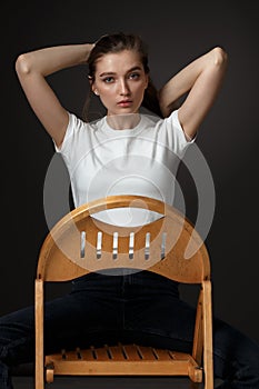 Charming brunette girl with long flowing hair dressed in white t-shirt and jeans poses sitting on the wooden chair on