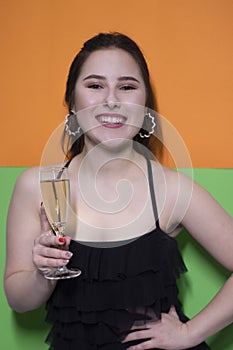 Charming brunette woman in black dress holding glass of champagne with an engagement ring inside on bright green and orange