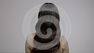 Charming brunette woman shaking her long wavy hair on a gray background. Back view