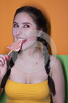 Charming brunette woman in yellow top holding red big lollipop on bright green and orange background. sweets concept. candy