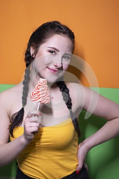 Charming brunette woman in yellow top holding red big lollipop on bright green and orange background. sweets concept. candy