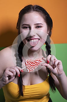 Charming brunette woman in yellow top holding red big lollipop on bright green and orange background. sweets concept. candy