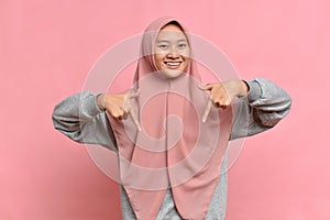 Charming carefree Muslim Female with positive expression, points down with both index fingers, wears hijab, isolated over pink